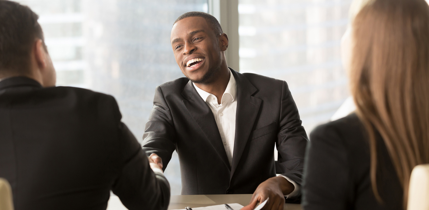 nojs Three people sat at a desk in an interview scenario with two men shaking hands and smiling.