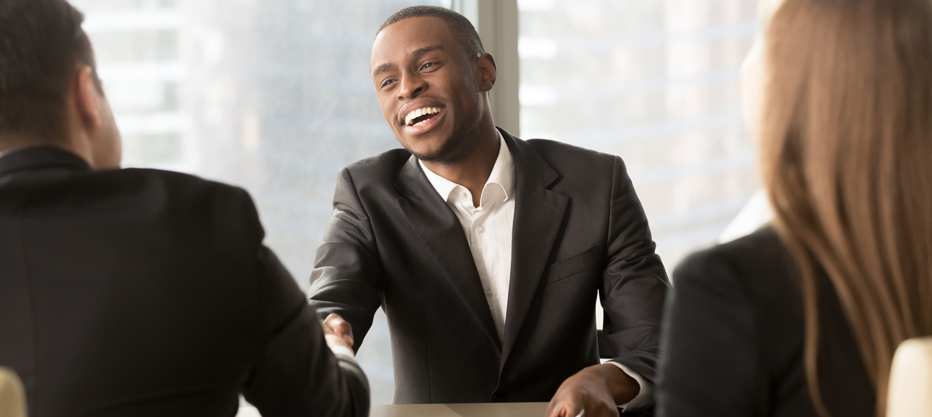 nojs Three people sat at a desk in an interview scenario with two men shaking hands and smiling.