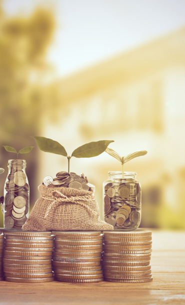 Five equal stacks of coins topped with glass jars and a hessian bag with more coins inside and plant sprouts representing growth.