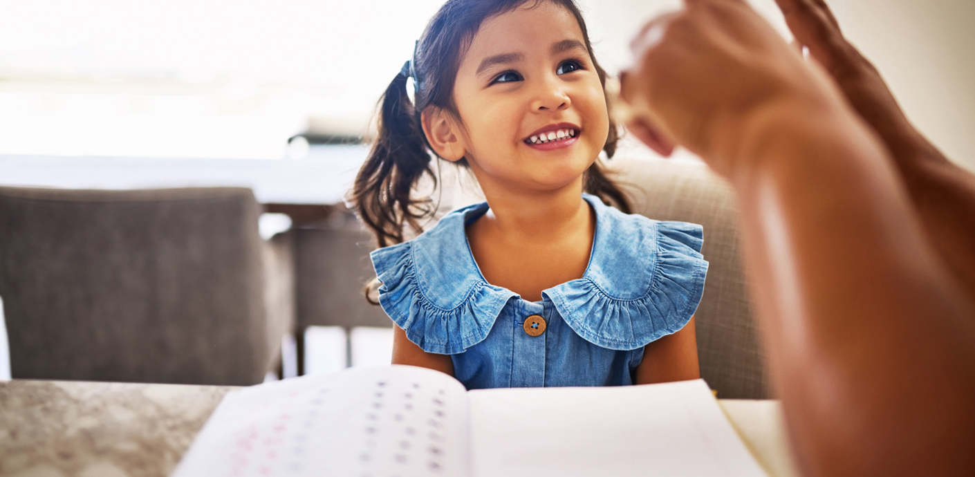 nojs A smiling girl sat at a table being taught maths.