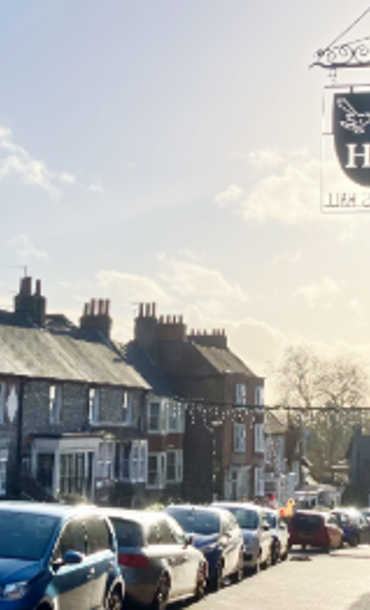 Traditional village high street with parked cars, historic buildings and a hanging pub sign on a sunny day.